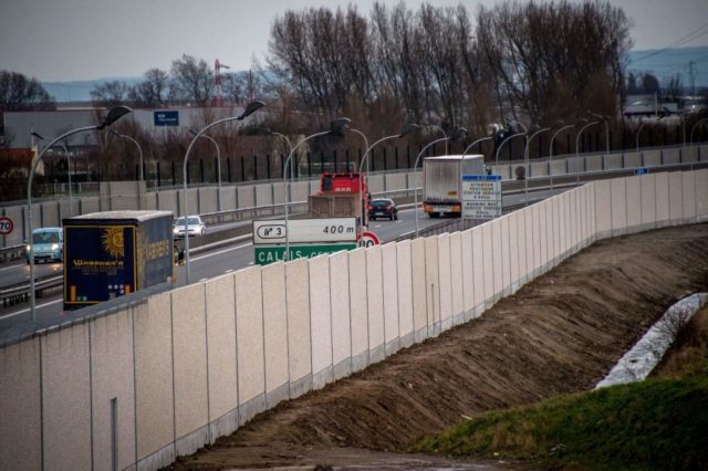 A photo taken on February 2, 2017 in Calais, shows trucks on a road next to a four-metre-high (13-foot) wall, running along a kilometre-long stretch of the main road leading to Calais port, aimed at stopping migrants who attempt to reach its shores. The wall was built to boost a network of wire fences that had failed to prevent near nightly attempts by migrants to waylay trucks en route to Europe's second-busiest port. Calais has for years been a staging post for attempts by migrants to sneak into Britain by stowing away on trucks or trains crossing the Channel. / AFP / PHILIPPE HUGUEN (Photo credit should read PHILIPPE HUGUEN/AFP/Getty Images)