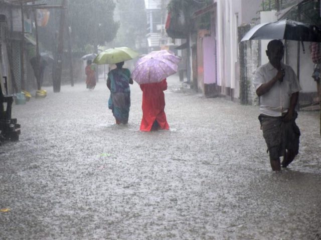 Menschen mit Regenschirmen waten indischen Balurghat durch eine überflutete Straße. Foto: Balurghat-Majidur Sardar/dpa