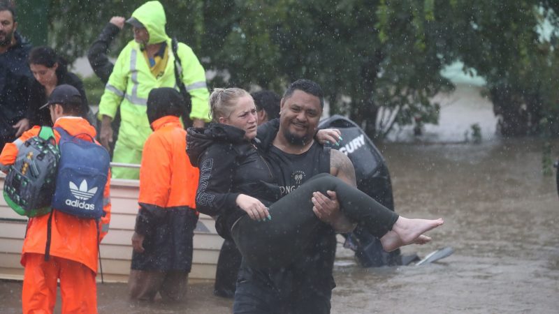Ein Mann trägt eine Frau durch das Hochwasser einer überschwemmten Straße im australischen Lismore.