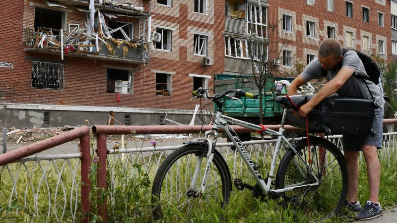 Ein Mann in Slowjansk zurrt einen Koffer auf seinem Fahrrad fest. Das Wohnhaus hinter ihm wurde bei einem nächtlichen Raketeneinschlag beschädigt.