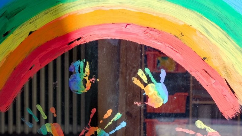 Kinderhände und ein Regenbogen am Fenster einer Kita.