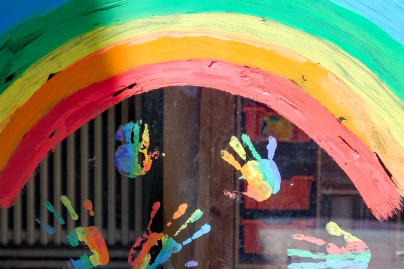 Kinderhände und ein Regenbogen am Fenster einer Kita.