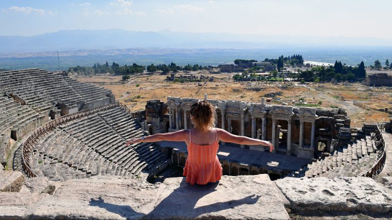 Eine Touristin breitet die Arme aus während sie auf den Ruinen der antiken Stadt und UNESCO-Weltkulturerbe Hierapolis sitzt.