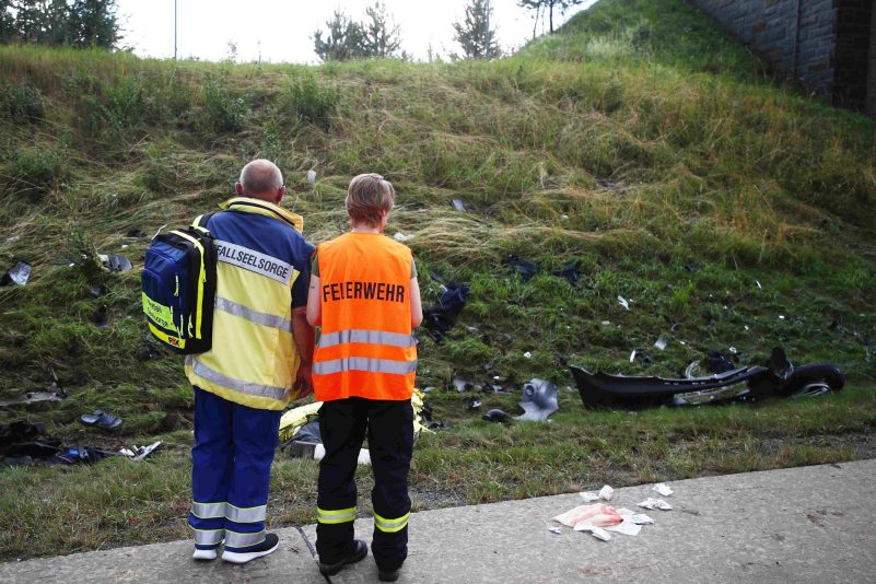 Ein Seelsorger und ein Mitglied der Feuerwehr an einer Unfallstelle auf der Autobahn 9 vor einer Böschung, die mit Trümmerteilen übersäht ist.