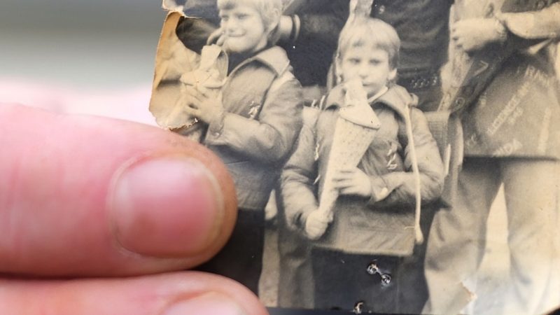 Kai Oppermann, in DDR-Kinderheimen groß geworden, zeigt ein Foto von seiner Einschulung. In der Bildmitte ist er in der vorderen Reihe mit Zuckertüte zu sehen. Links daneben steht sein Bruder Patrick.