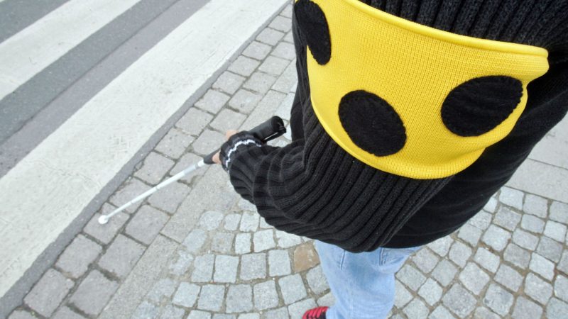 Ein blinder Mann geht mit seinem weißen Taststock und einer Armbinde mit drei Punkten über eine Straße in Hannover.
