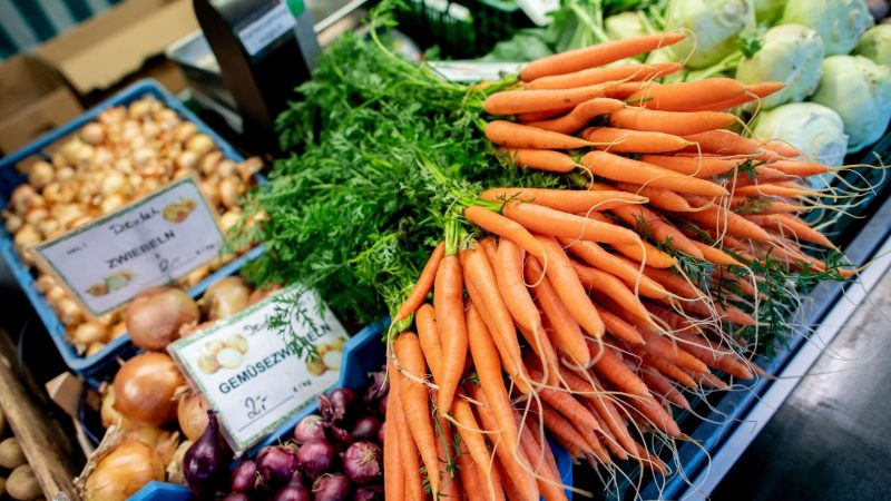 Karotten und Zwiebeln an einem Obst- und Gemüsestand auf einem Wochenmarkt. Die Preise für Nahrungsmittel bleiben weiter hoch.