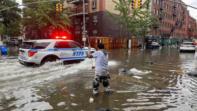 Hochwasser auf den Straßen von New York: Im Stadtteil Brooklyn versucht ein Mann, mit einem  Stock einen Abfluss zu reinigen, während ein Polizeiwagen vorbeifährt.