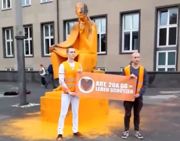 Zwei Mitglieder der „Letzten Generation“ posieren vor der mit Farbe überzogenen Albertus-Magnus-Statue vor der Kölner Uni.