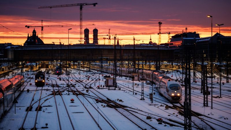 Der Tag beginnt: Die Sonne geht über den Gleisanlagen vor dem Münchner Hauptbahnhof und der Frauenkirche auf und taucht den Himmel in bunte Farben.