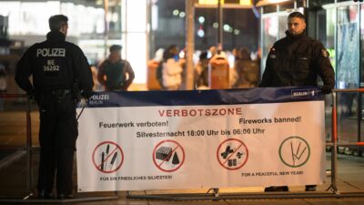 Polizeibeamte stehen an einem Plakat, das auf die Böllerverbotszone am Berliner Alexanderplatz hinweist.