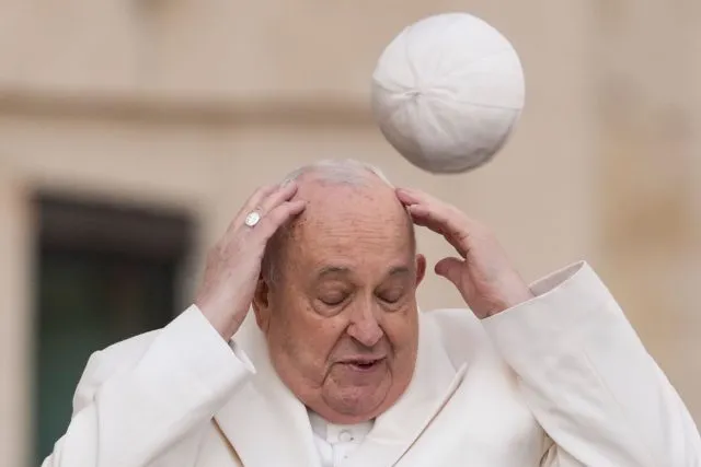 Bei seiner Generalaudienz auf dem Petersplatz versucht Papst Franziskus, seinen Pileolus aufzufangen, den der Wind weggeweht hat.
