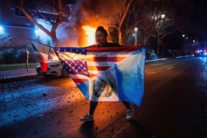 Eine Frau mit mehreren Flaggen geht an einem brennenden Auto vorbei, während sie gegen die Razzien der Trump-Regierung gegen Einwanderer in Los Angeles protestiert.