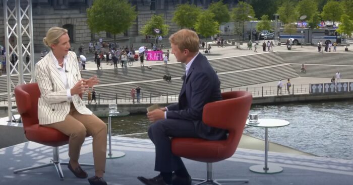 Das Bild zeigt Alice Weidel und ARD-Hauptstadtstudioleiter Markus Preiss während des ARD-Sommerinterviews vom 20. Juli 2025. Im Hintergrund rechts sind ein Polizeiwagen und der Aktivistenbus des „Zentrums für politische Schönheit“ (ZPS) zu sehen. Foto: Bildschirmfoto/ARD-Mediathek