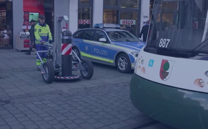 Ein Mitarbeiter einer privaten Sicherheitsfirma wartet darauf, dass er nach der Durchfahrt der Straßenbahn am Augsburger Christkindlesmarkt einen mobilen Schutzpoller wieder auf das Gleis schieben kann. Foto: Bildschirmfoto/BR