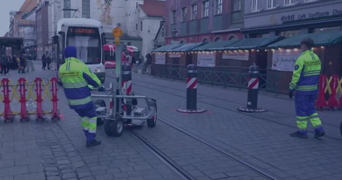Mitarbeiter einer privaten Sicherheitsfirma räumen einen mobilen Schutzpoller aus dem Weg, damit eine Straßenbahn die Augsburger Maximilianstraße in unmittelbarer Nähe des Christkindlesmarktes passieren kann. Foto: Bildschirmfoto/BR