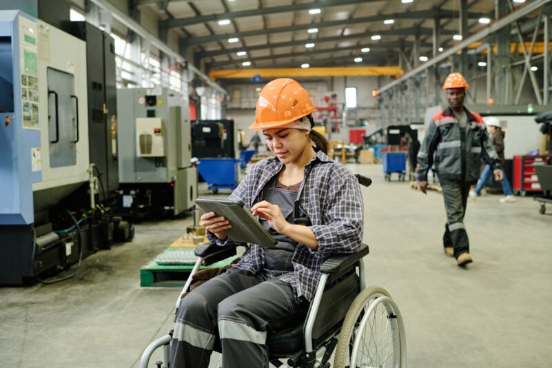 Worker in wheelchair using tablet for managing processes in modern industrial facility. Colleague in background walking between machines, both wearing safety gear. Foto: Pressmaster/iStock